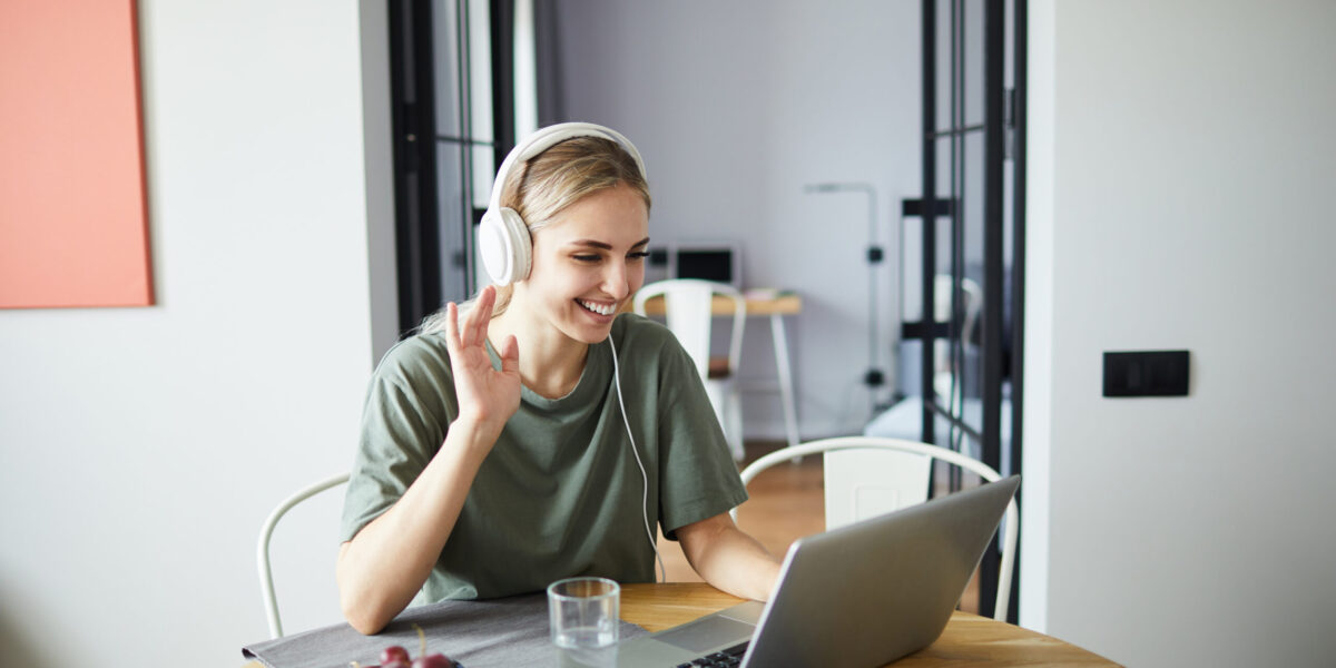 Cheerful girl in headphones waving her hand to friend while communicating through video-chat in front of laptop